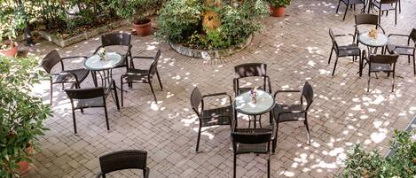 An outdoor patio area with multiple round tables and wicker chairs, surrounded by potted plants and trees.