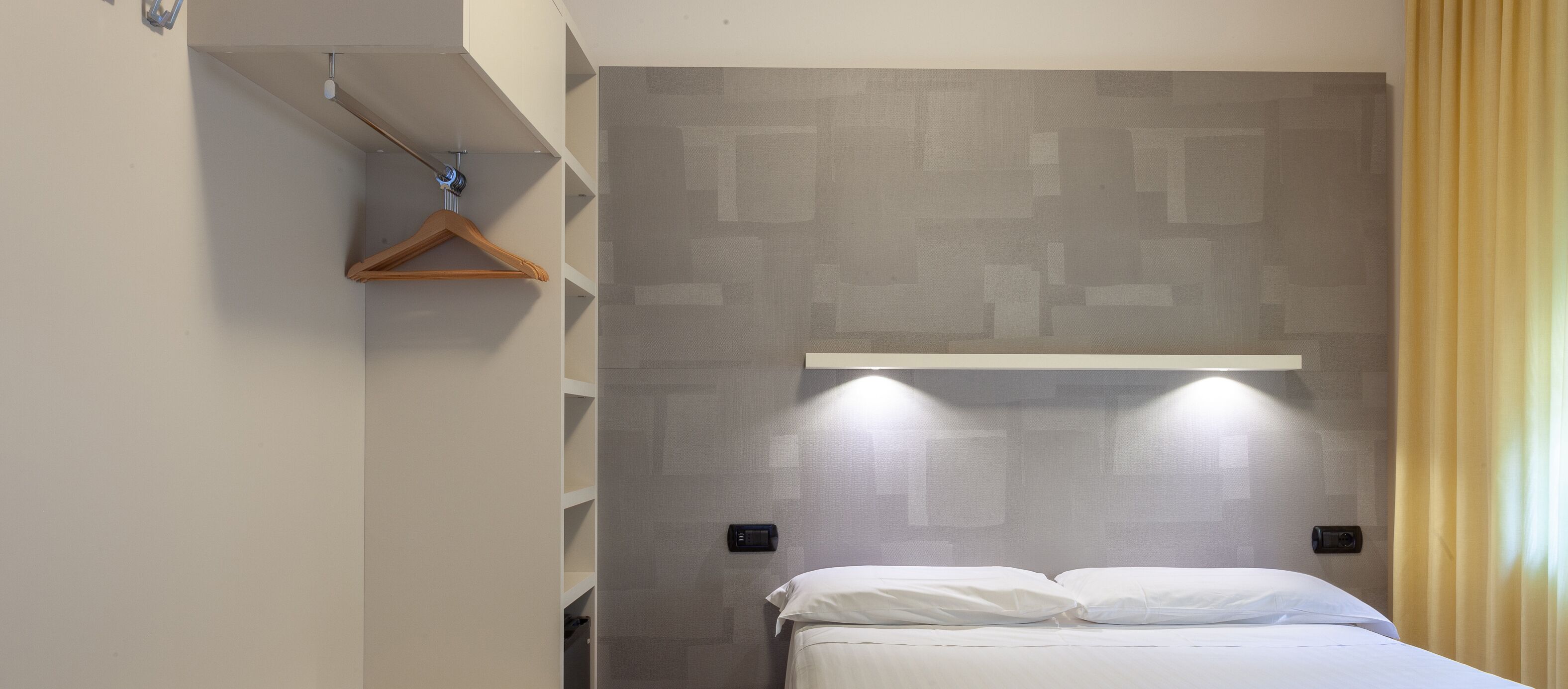 Una habitación de hotel con una cama, un perchero de madera, una lámpara de pared y un estante.