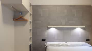 Una habitación de hotel con una cama, un perchero de madera, una lámpara de pared y un estante.