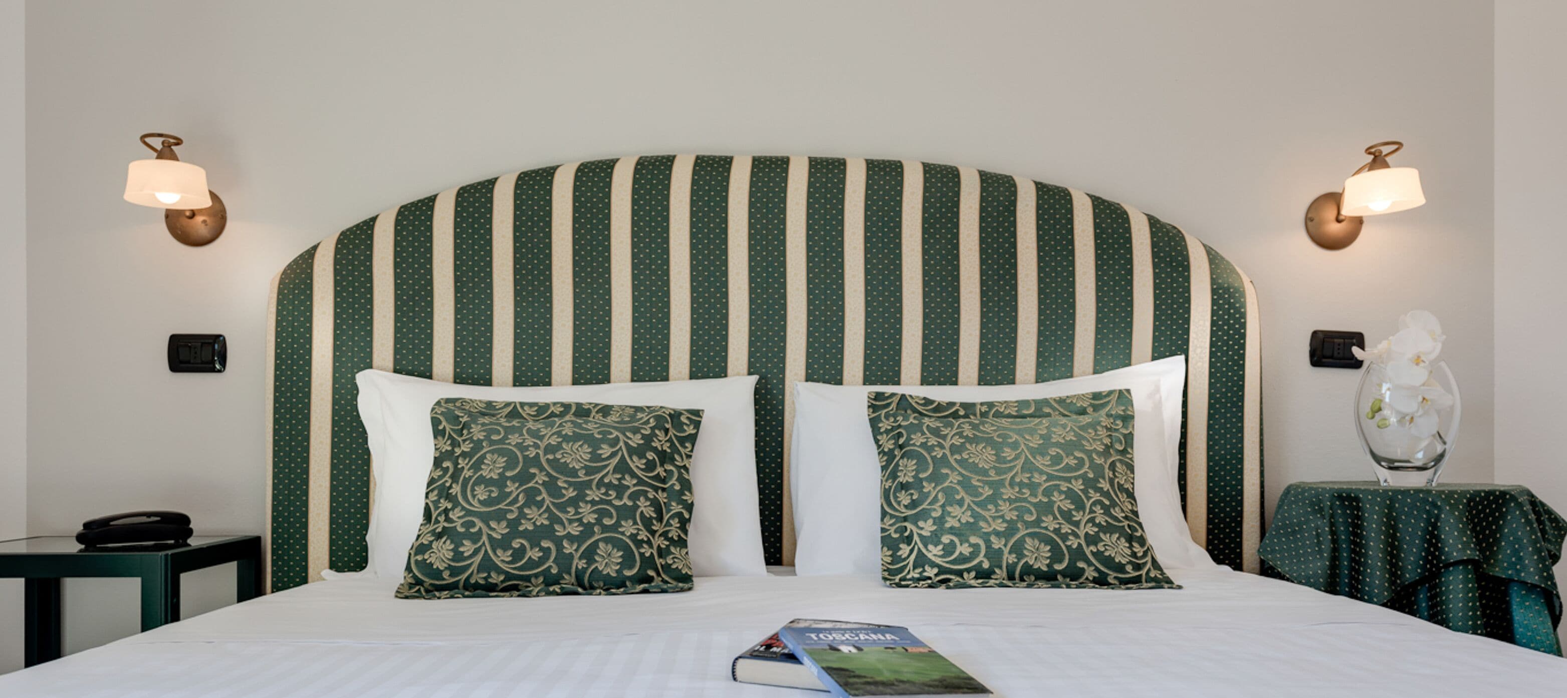 Habitación de hotel con una cama grande, dos almohadas, una mesita de noche con lámpara y una pequeña mesa con un jarrón de flores.