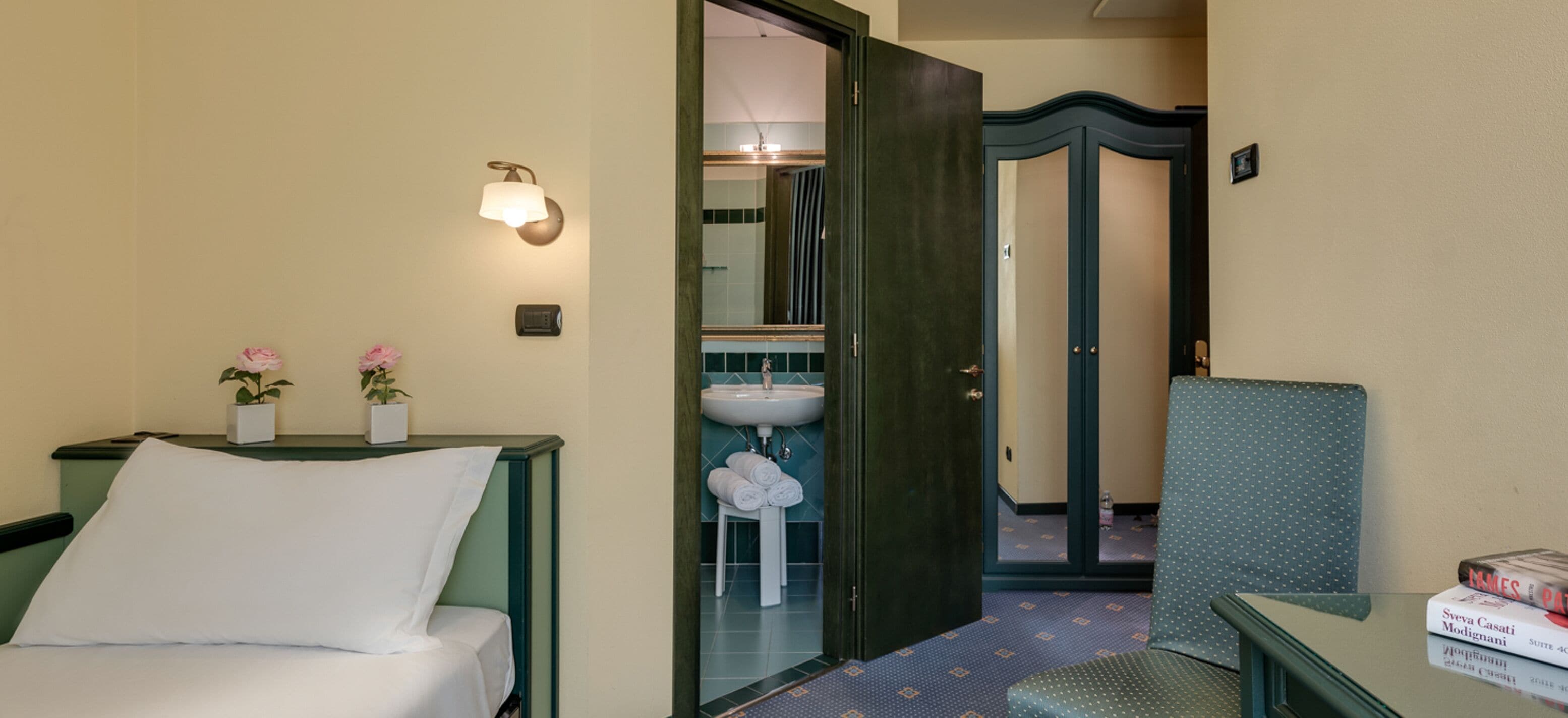 A hotel room with a bed, a bedside table with two potted plants, a nightstand, a small table with a book, and a bathroom visible through an open door.
