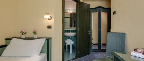 A hotel room with a bed, a bedside table with two potted plants, a nightstand, a small table with a book, and a bathroom visible through an open door.