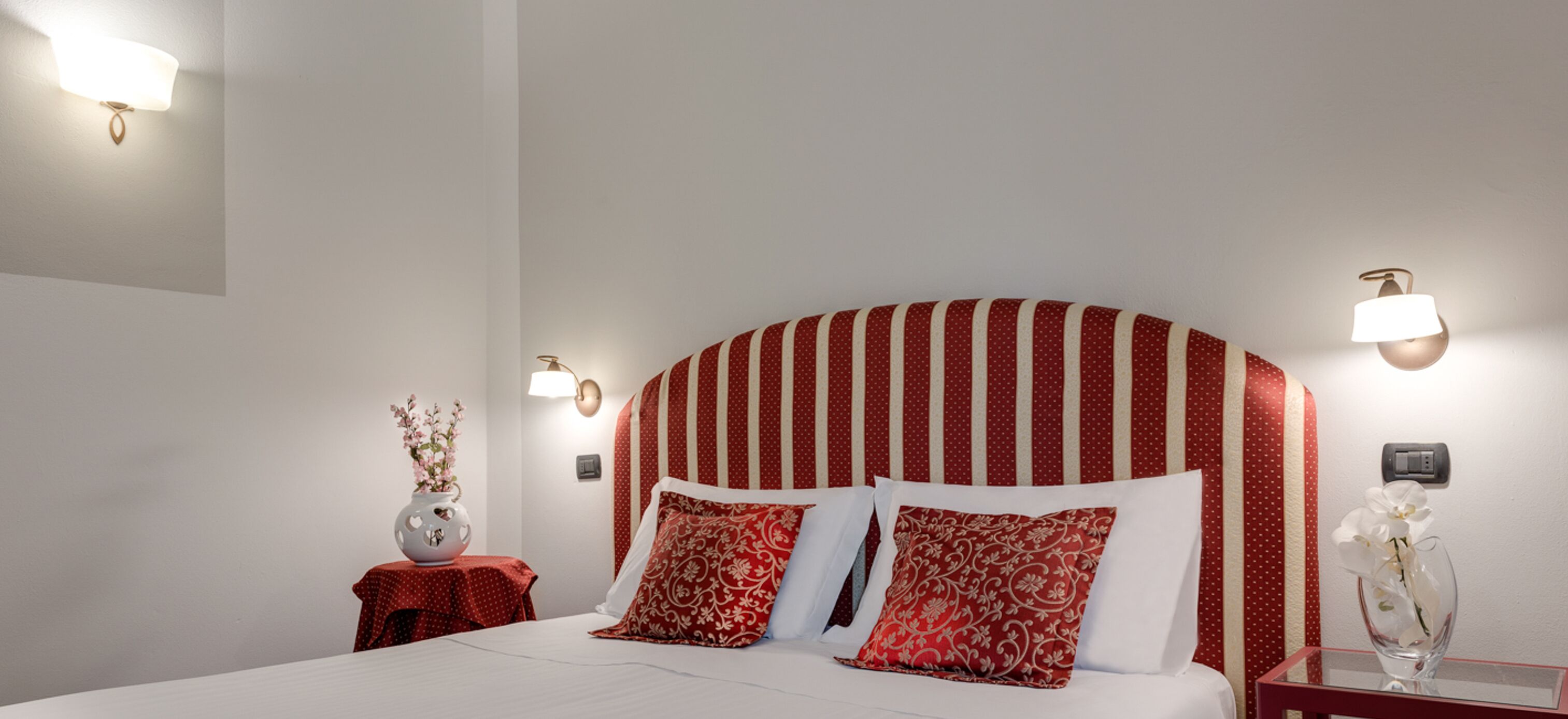 Une chambre d’hôtel avec un grand lit, une tête de lit à rayures rouges, deux tables de chevet avec des lampes et un vase rempli de fleurs posé sur l’une des tables.