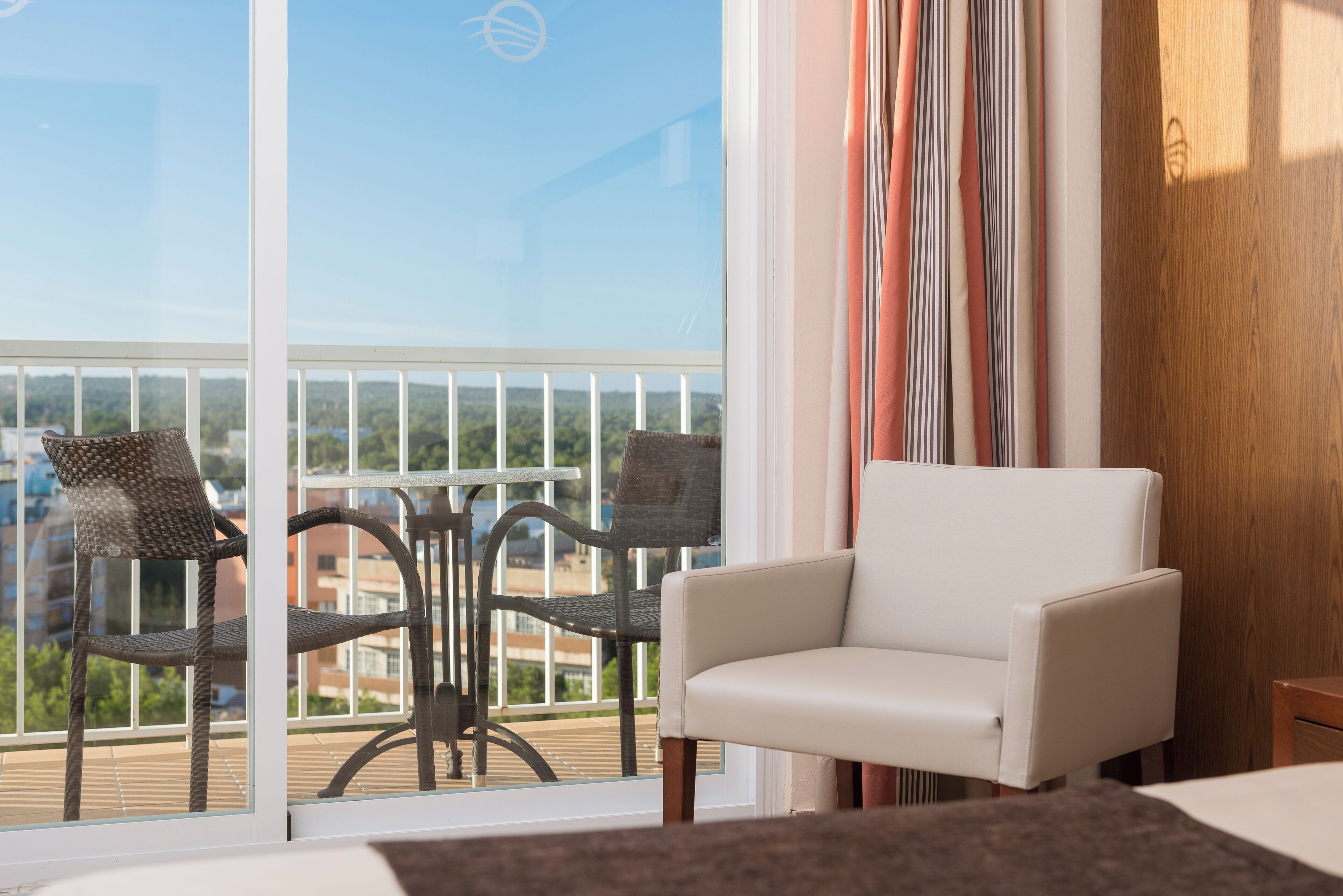 Un balcón con sillas de mimbre y un sillón blanco, con vista a un paisaje urbano.