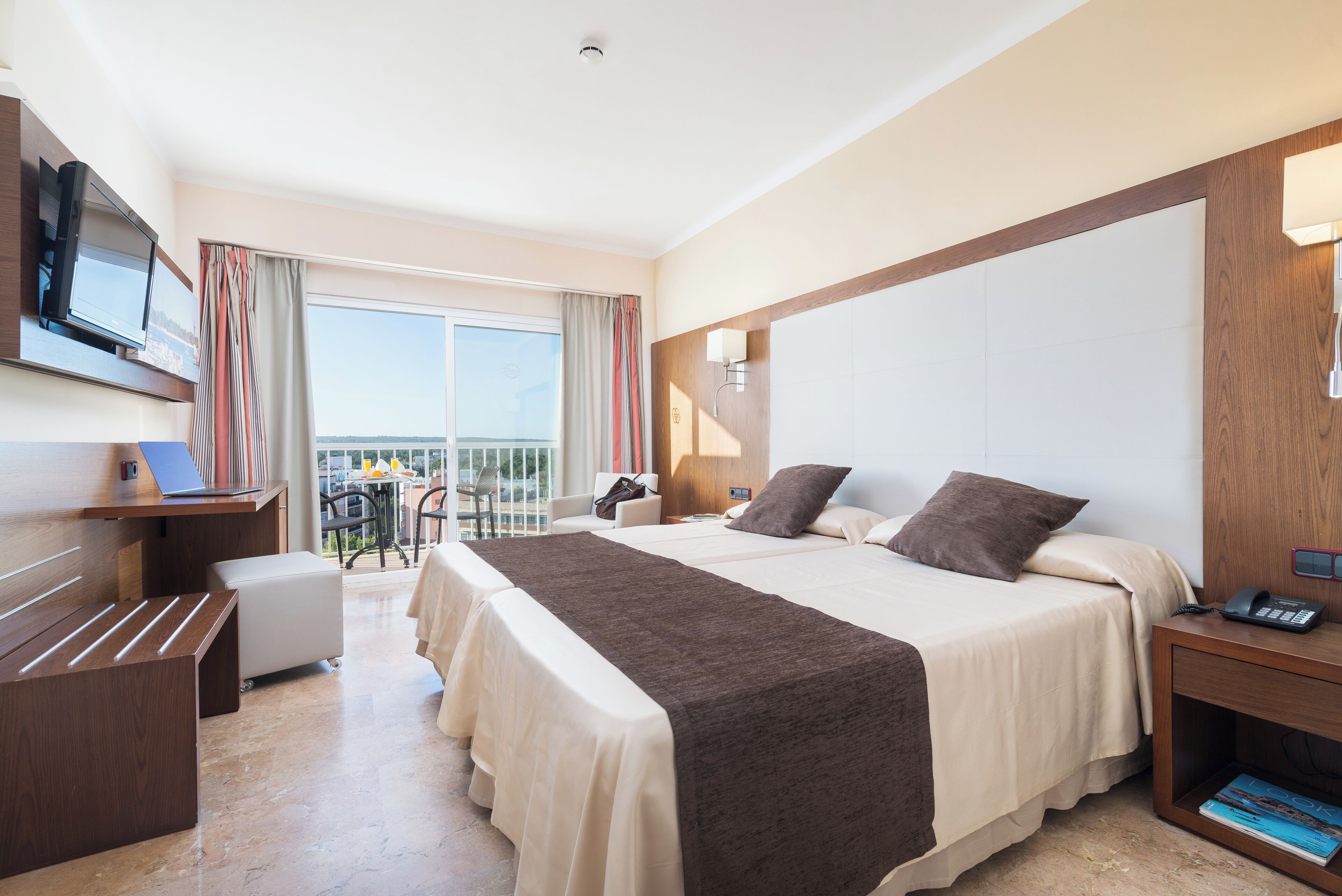 Habitación de hotel con una cama grande, un escritorio con una computadora portátil, un televisor y un balcón con vistas.