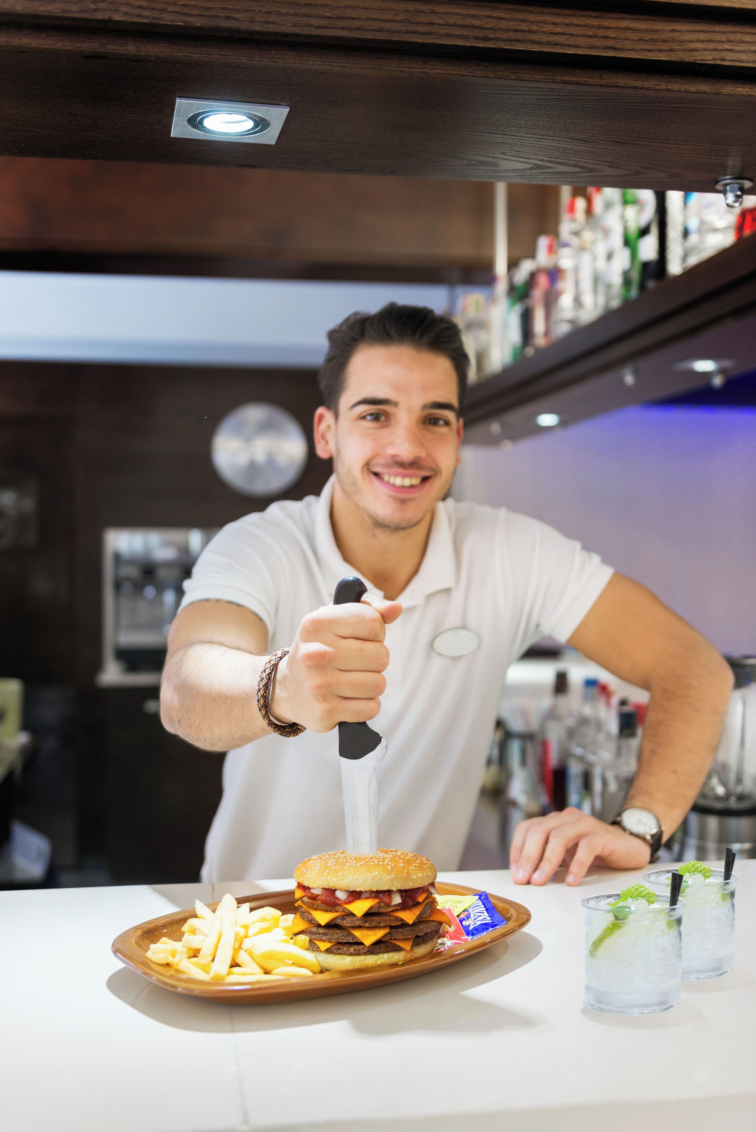 Una persona al bancone di un bar con un hamburger, patatine fritte e due bevande.