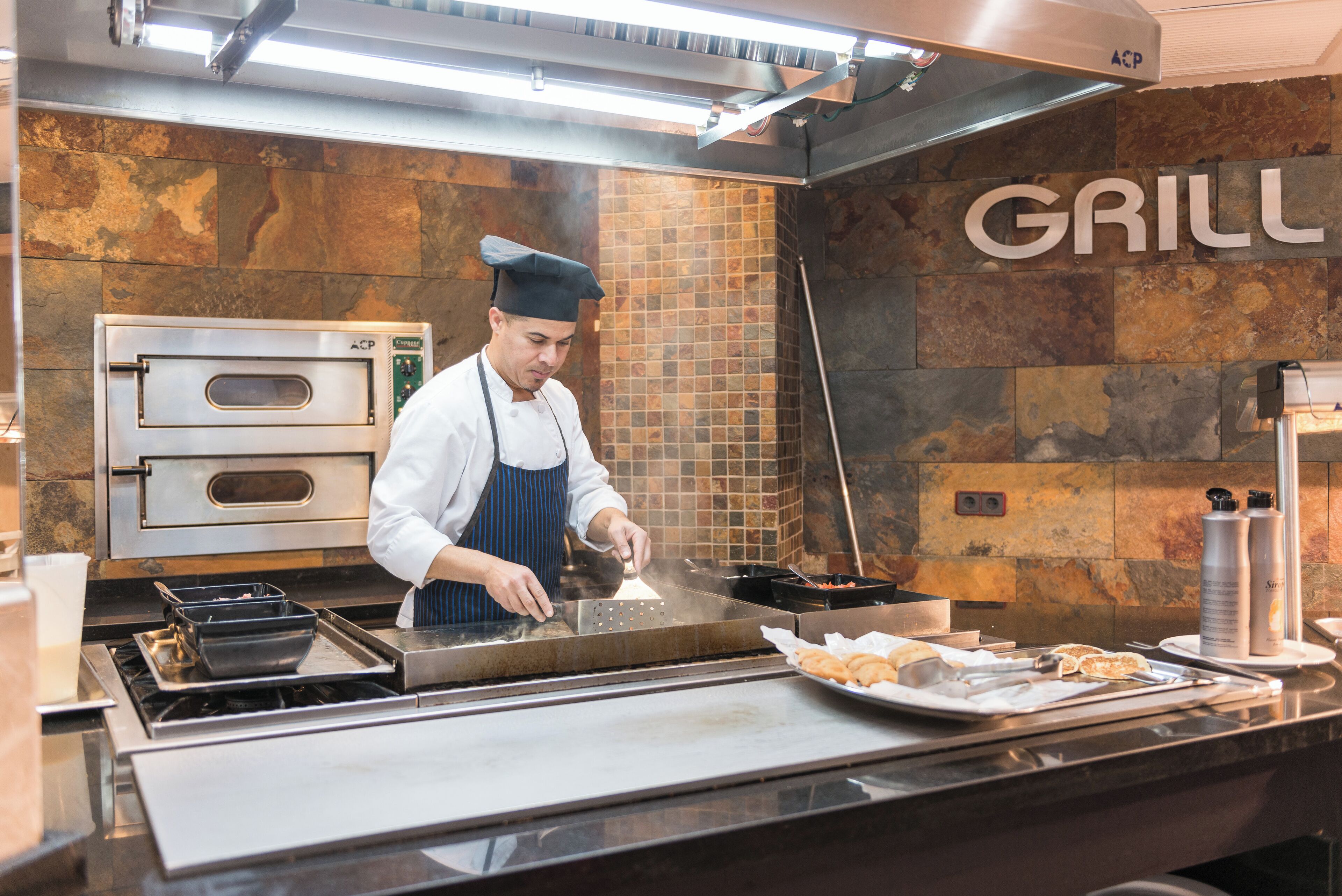 A chef cooking at a commercial kitchen with a stone wall and a 'GRILL' sign.