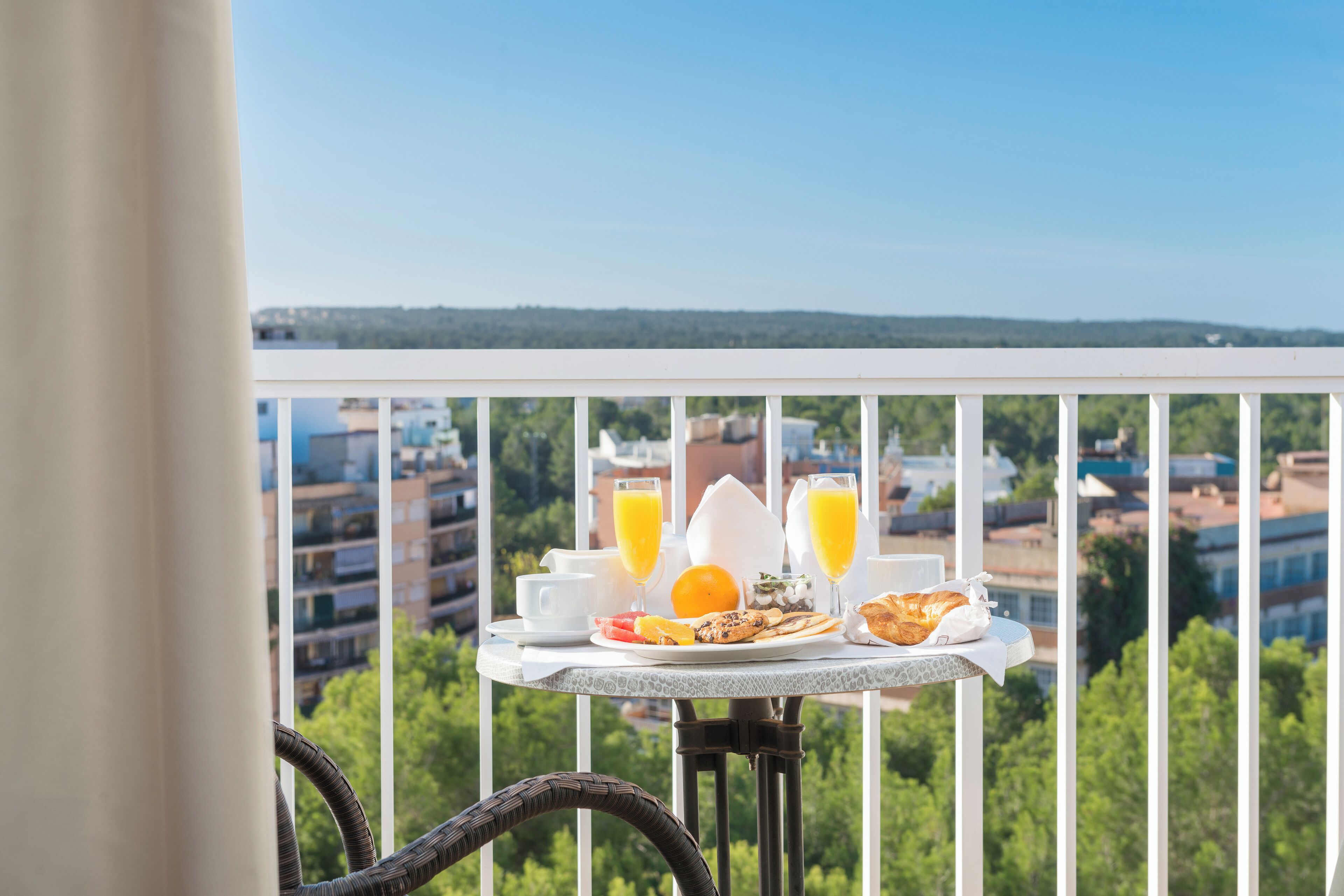 Mesa en balcón con alimentos para el desayuno y vista a un paisaje urbano.