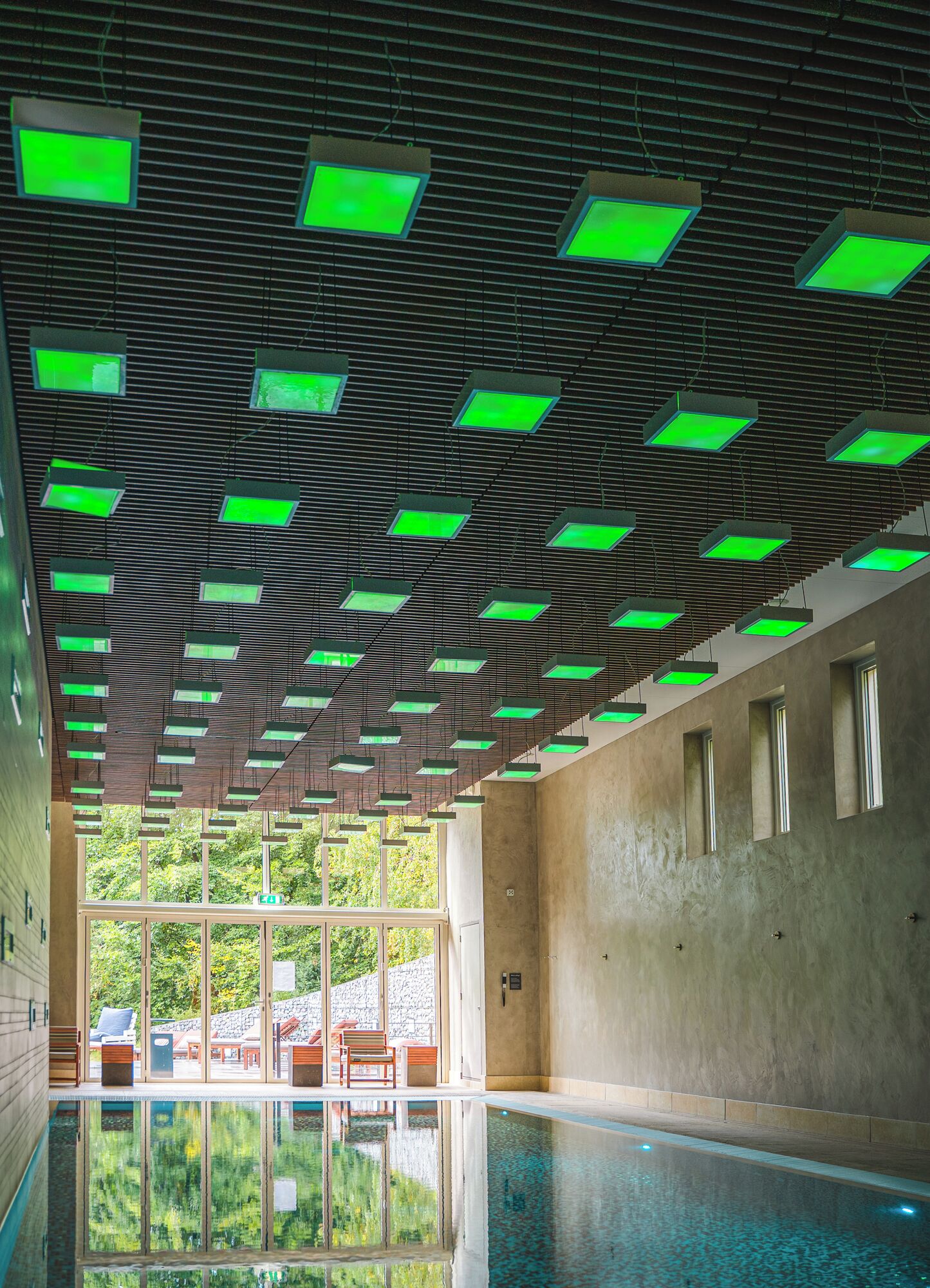 Une piscine intérieure moderne dotée d’un plafond composé de panneaux éclairés en vert, de grandes fenêtres et offrant une vue sur un espace extérieur aménagé avec des sièges.