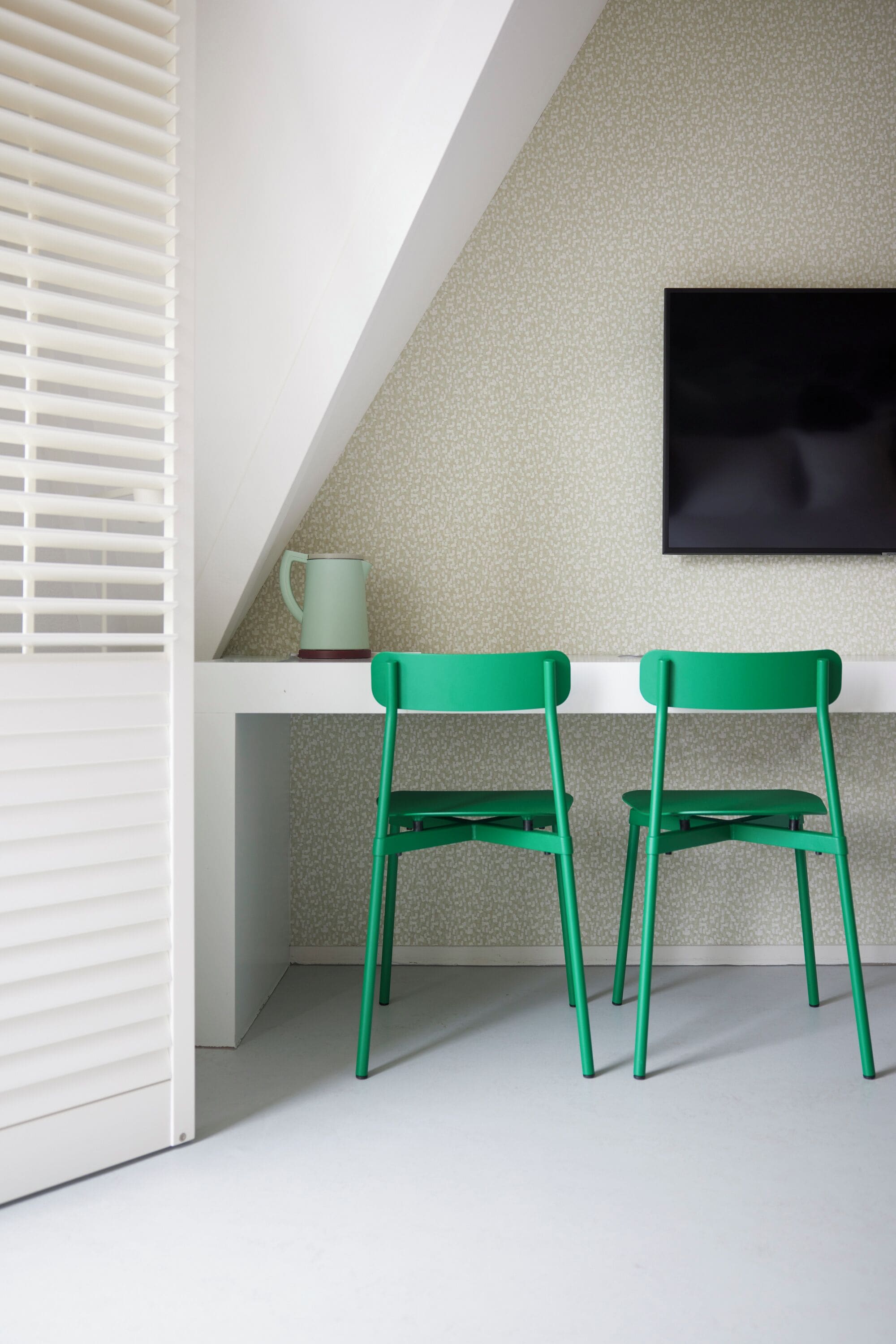 Ein Zimmer mit einem weißen Schreibtisch, zwei grünen Stühlen, einem weißen Fenster mit Jalousien und einer strukturierten Wand.