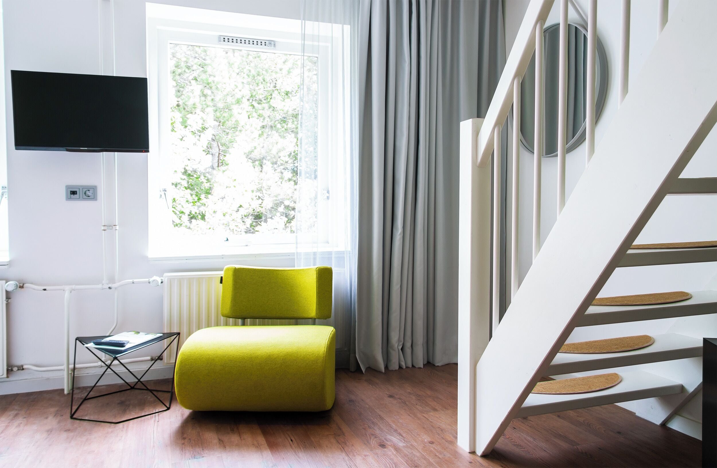 Una habitación moderna con suelo de madera, una silla amarilla, una mesa de vidrio, un televisor montado en la pared y una ventana con cortinas.