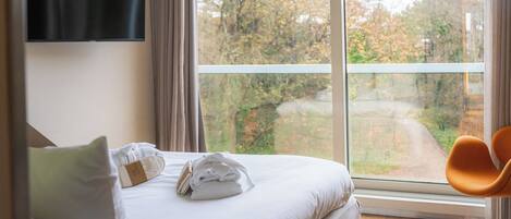 Quarto de hotel com cama, televisão, janela com vista para árvores e um caminho, e uma cadeira.