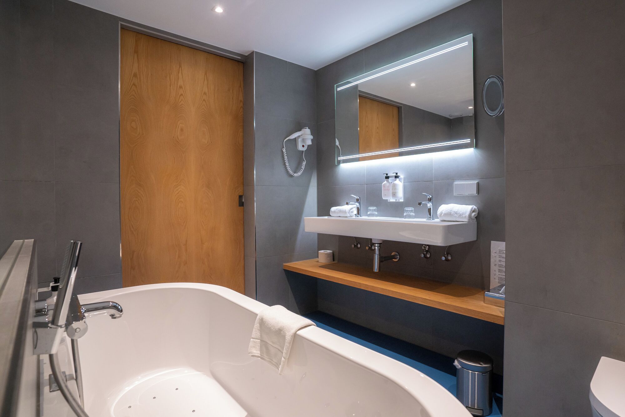 Un baño moderno con una bañera independiente, una puerta de madera, un espejo grande y un lavabo con toallero.