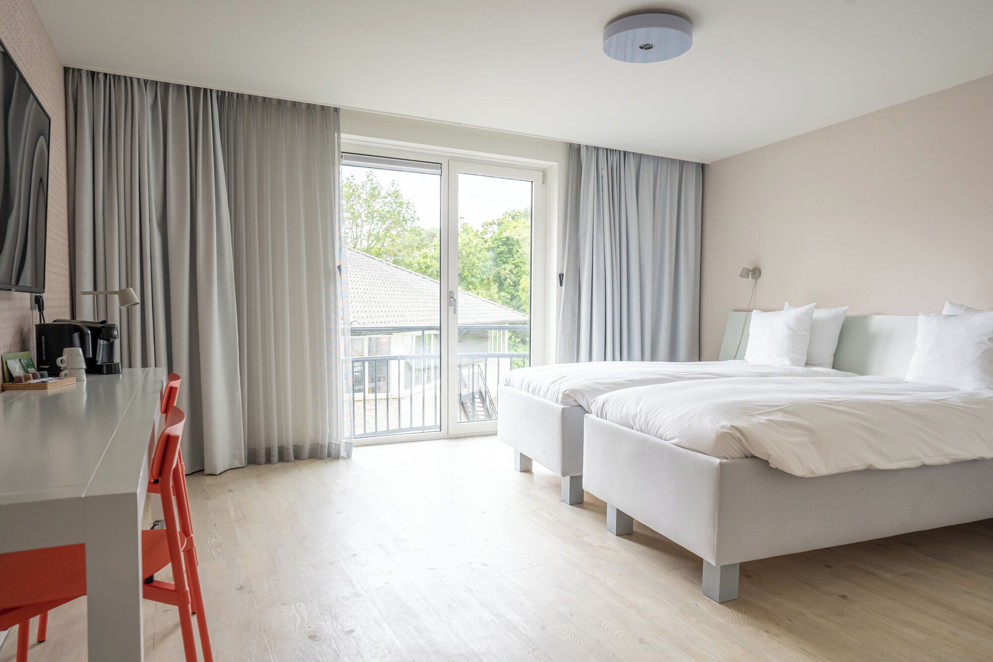 A modern hotel room with a bed, a desk with a coffee maker, and a view of greenery outside.
