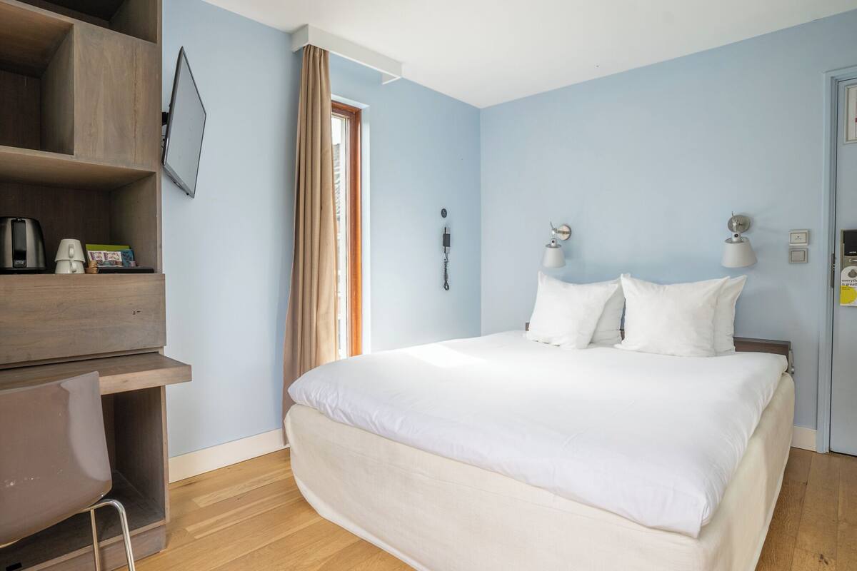 A hotel room with a bed, a desk, a chair, a telephone, and a door.