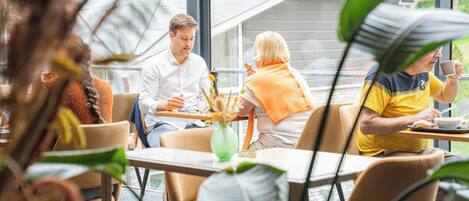Un café avec des tables et des chaises, de grandes fenêtres et des plantes d’intérieur.