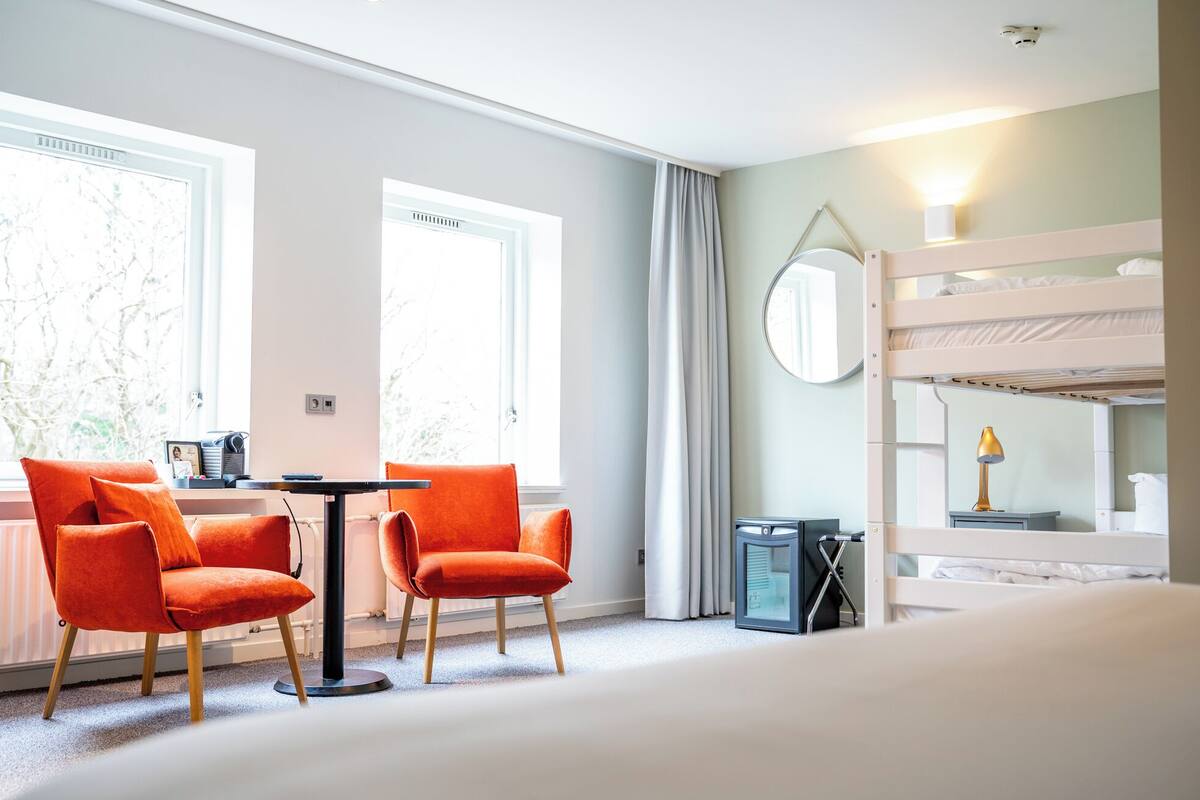 A hotel room with a bunk bed, a desk with two chairs, a mirror, and a window with curtains.