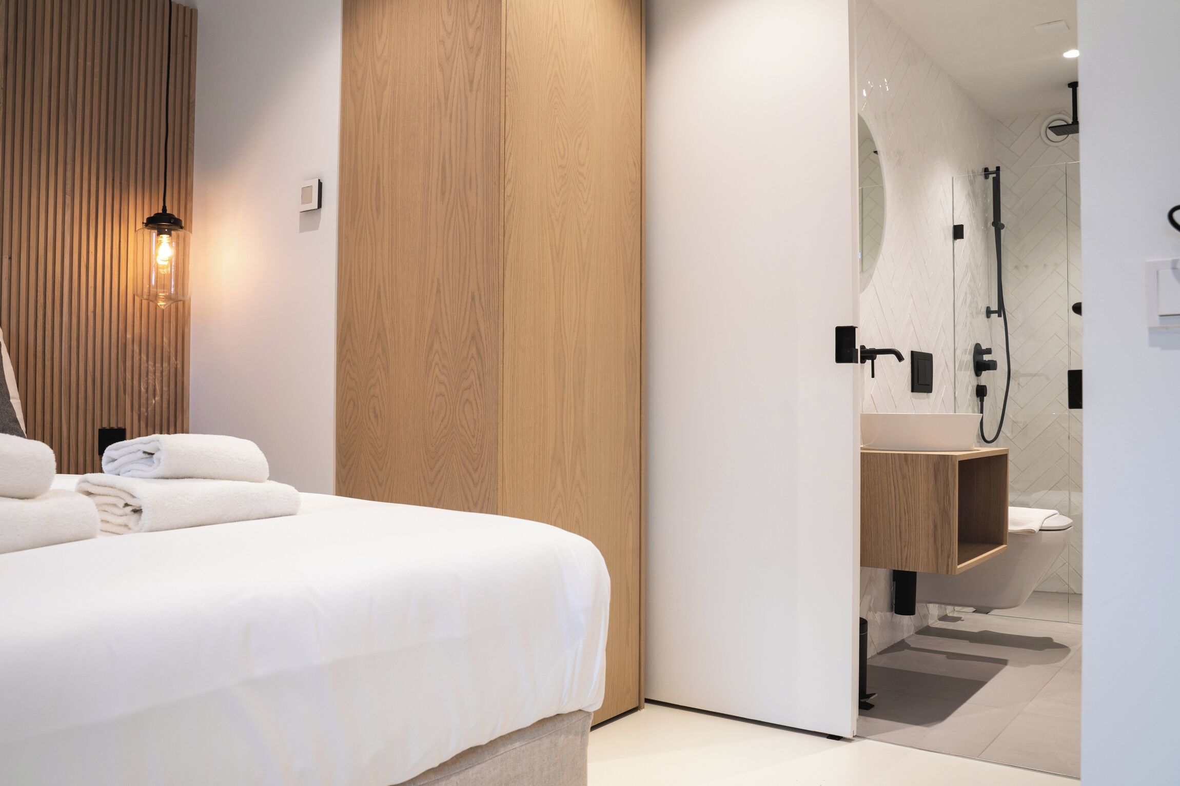 Une chambre d’hôtel moderne avec un lit soigneusement fait, un mur recouvert de panneaux en bois et une salle de bain équipée d’une douche, de toilettes et d’un lavabo.
