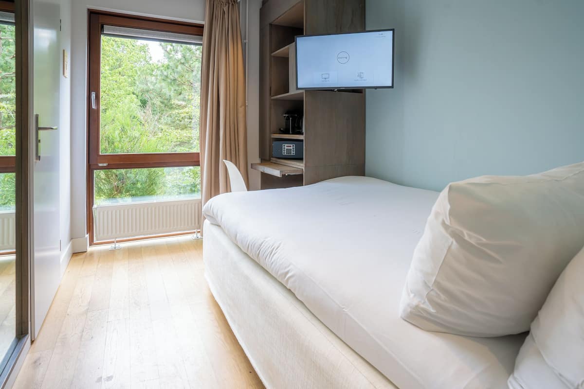 A bedroom with a bed, a television mounted on the wall, a window with curtains, and a view of greenery outside.