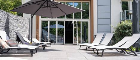Un área de descanso exterior moderna con sillones reclinables y un gran paraguas, que da paso a la entrada de un edificio.