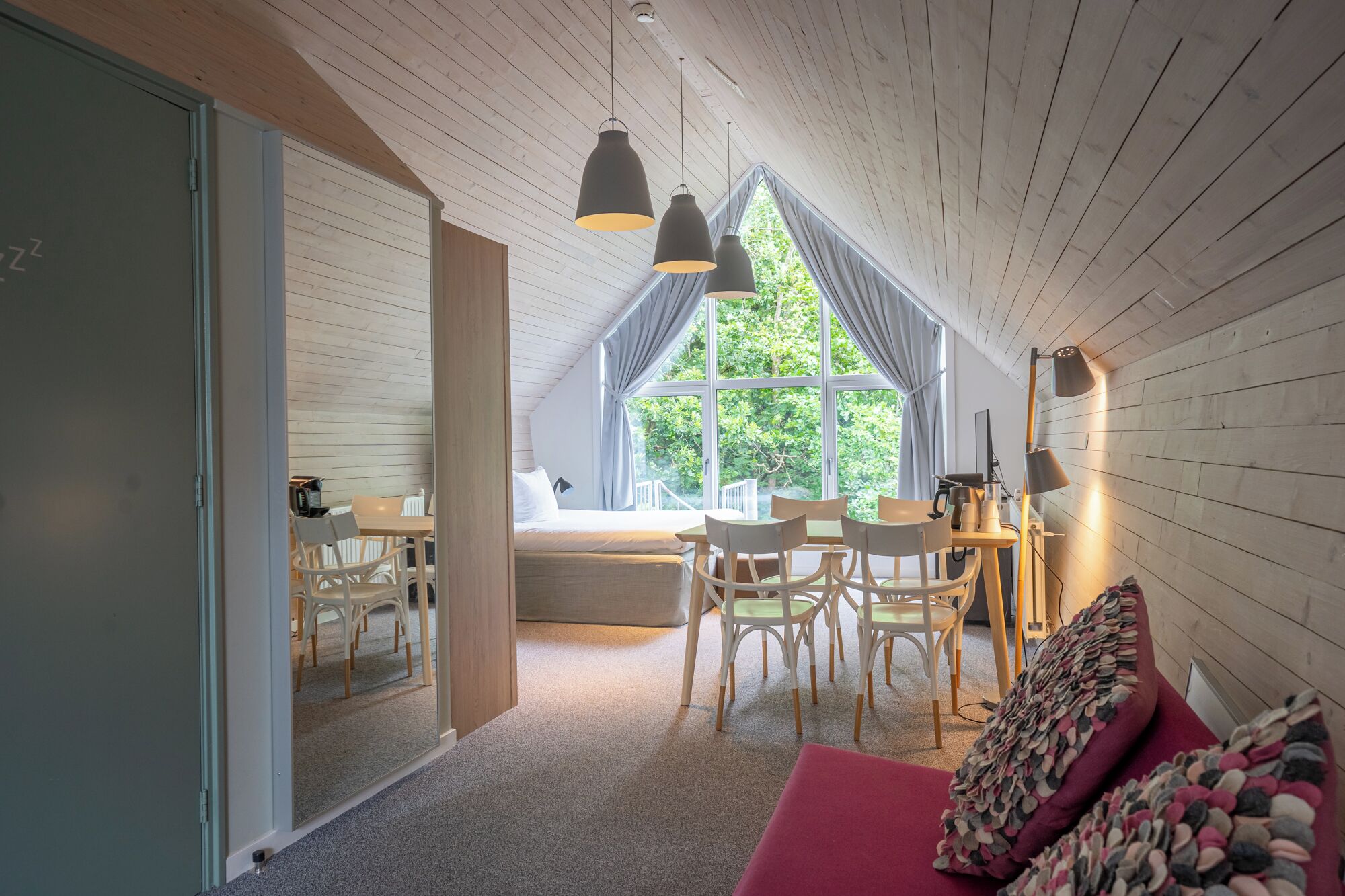 A cozy room with a wooden ceiling, a large window with curtains, a dining table with chairs, a bed, and a red sofa.