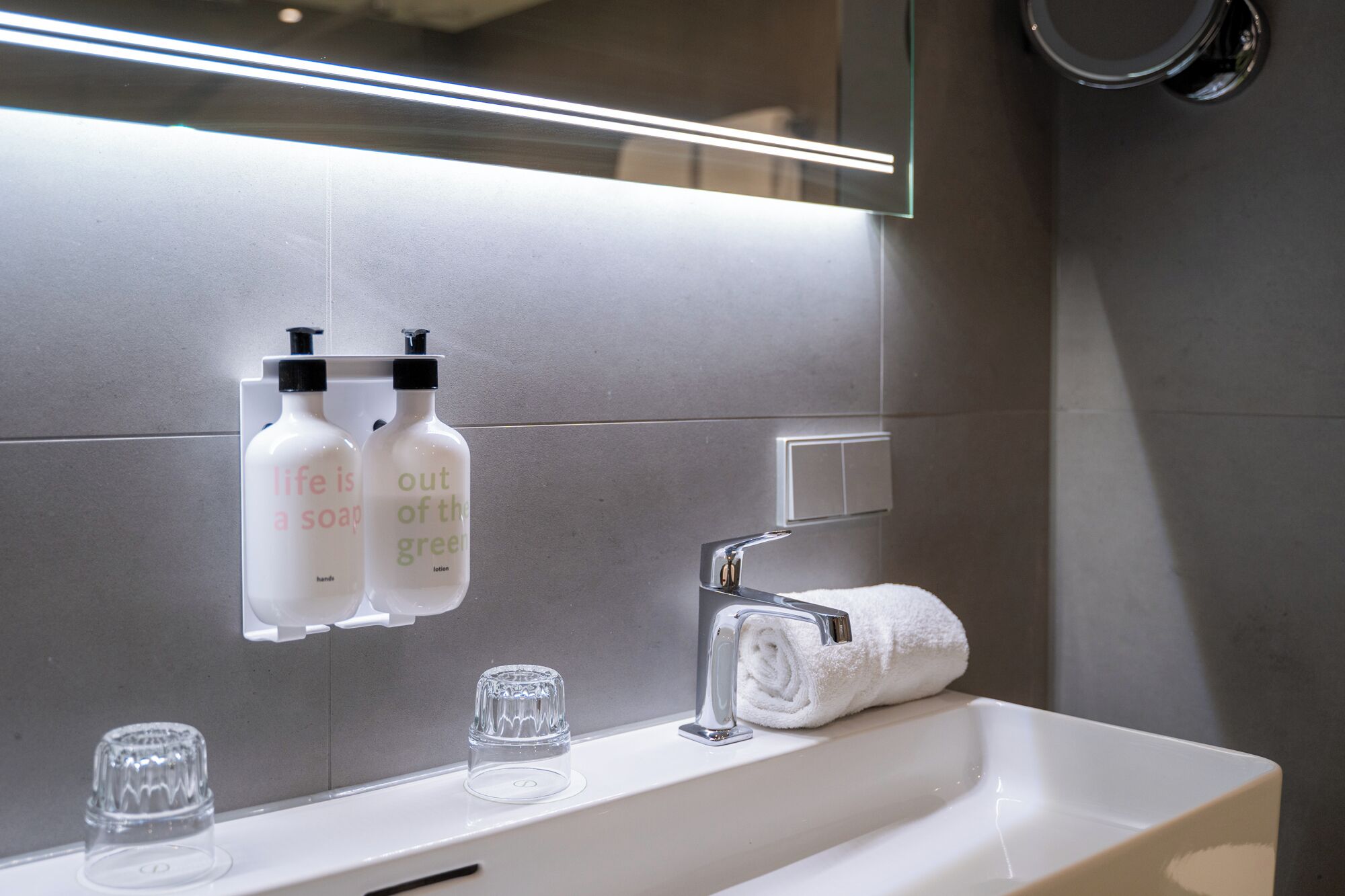 A modern bathroom with a white sink, two soap dispensers labeled 'life is a soap' and 'out of the green', a towel, and a glass soap dish.