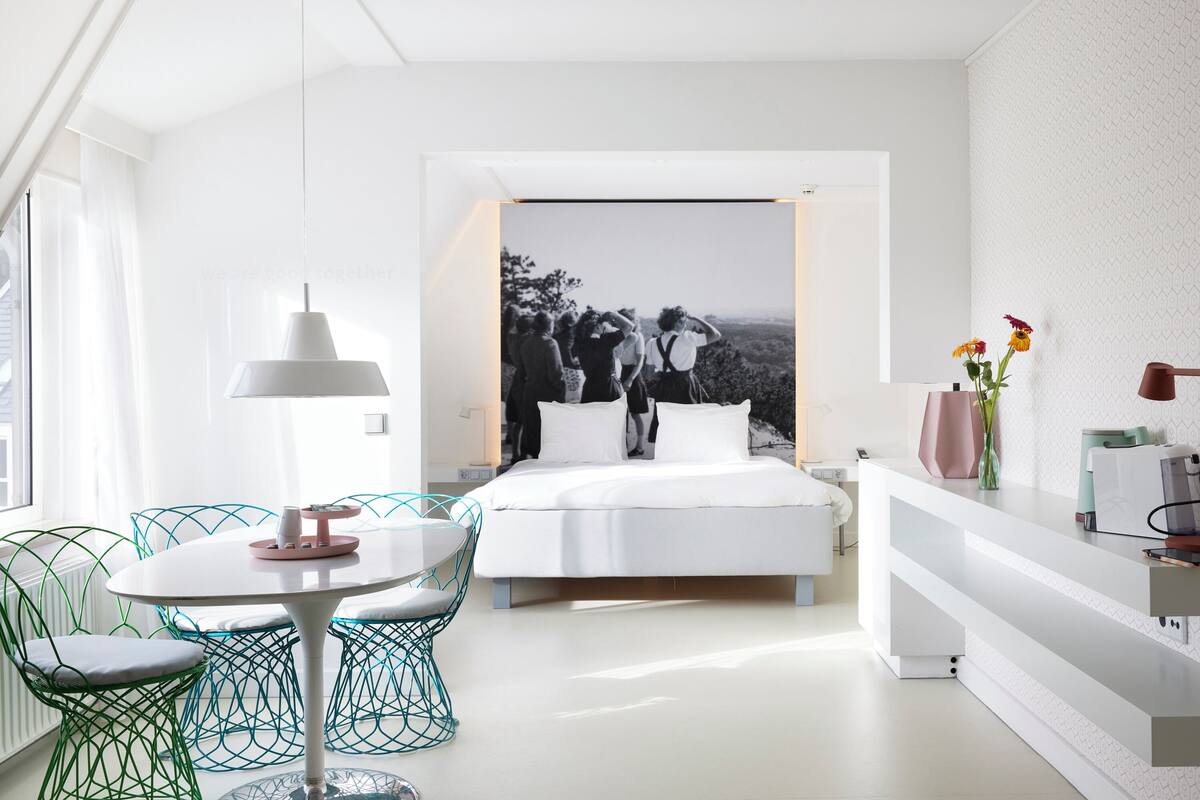 A modern hotel room with a bed, a dining table with chairs, a desk, and a large window.