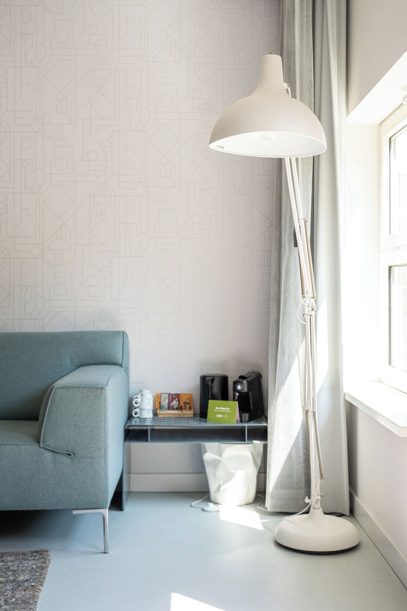 Una habitación moderna con un sofá gris, una lámpara de pie blanca, una máquina de café y una pequeña mesa con un cartel verde.