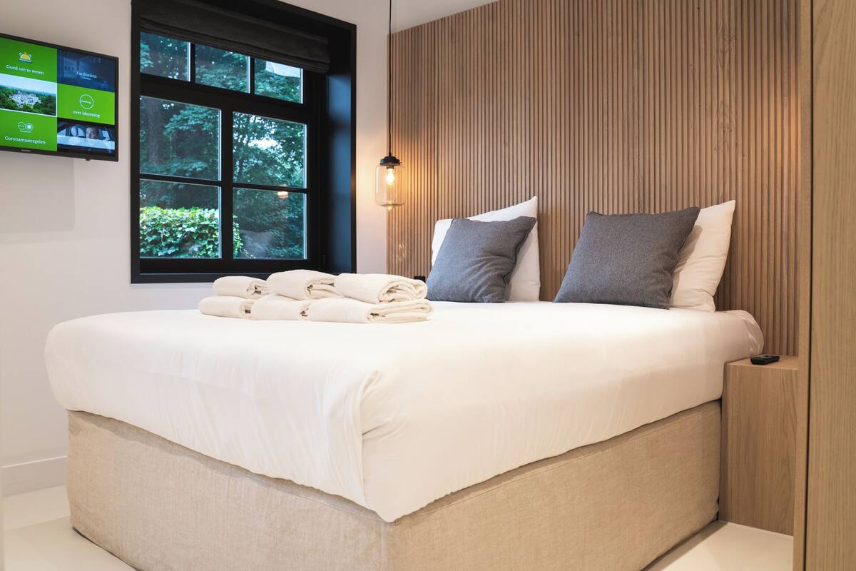 A neatly made bed with pillows and folded towels, a wooden headboard, and a television mounted on the wall.
