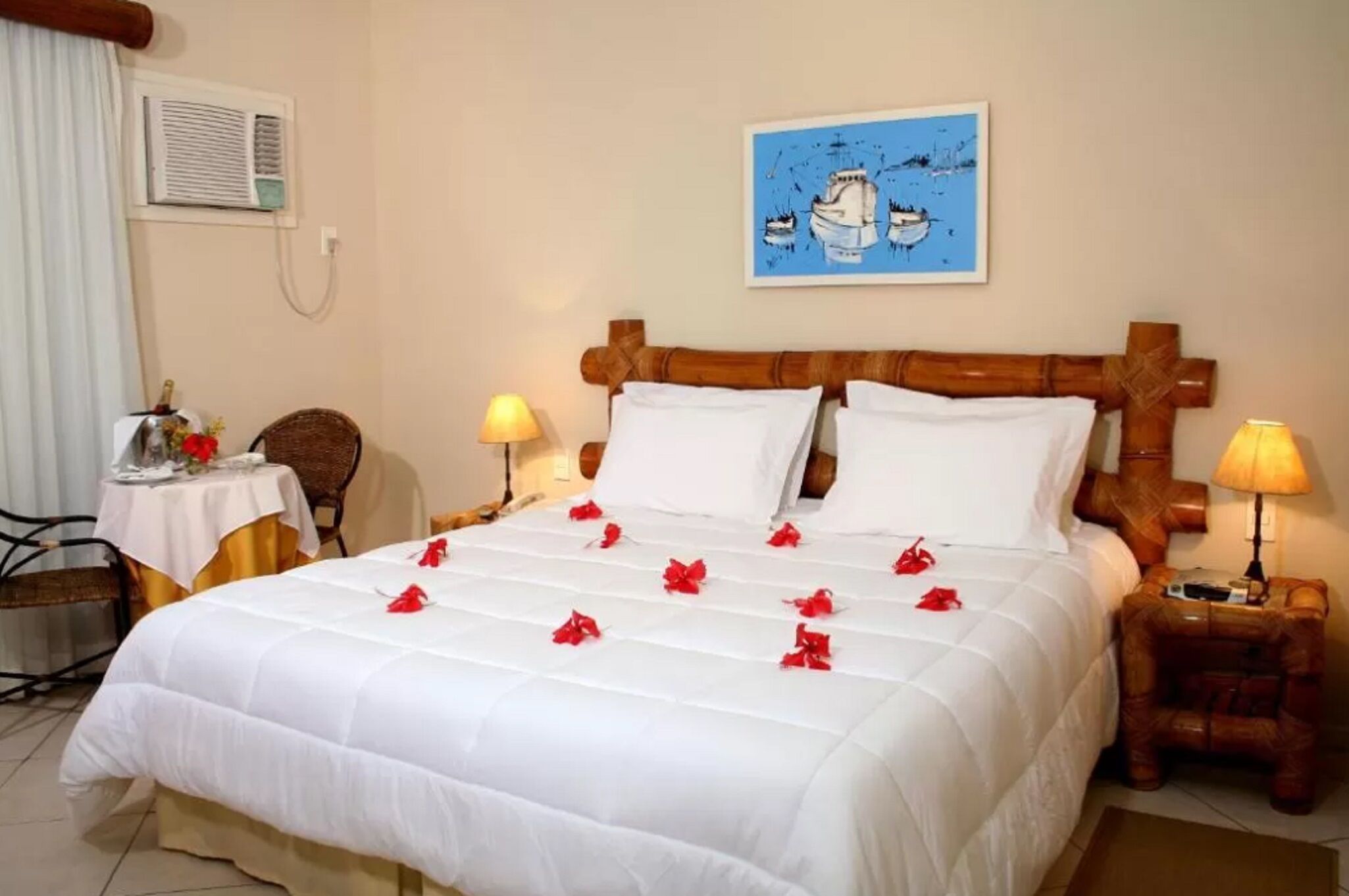 A neatly made bed with white bedding and red flowers, a wooden headboard, a small table with a lamp, and a chair.