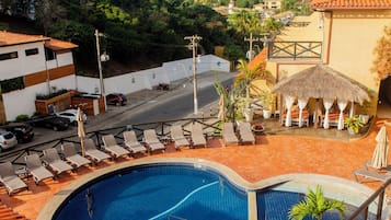 Una piscina en la azotea con diseño curvo, rodeada de sillones de descanso y una estructura con techo de paja.