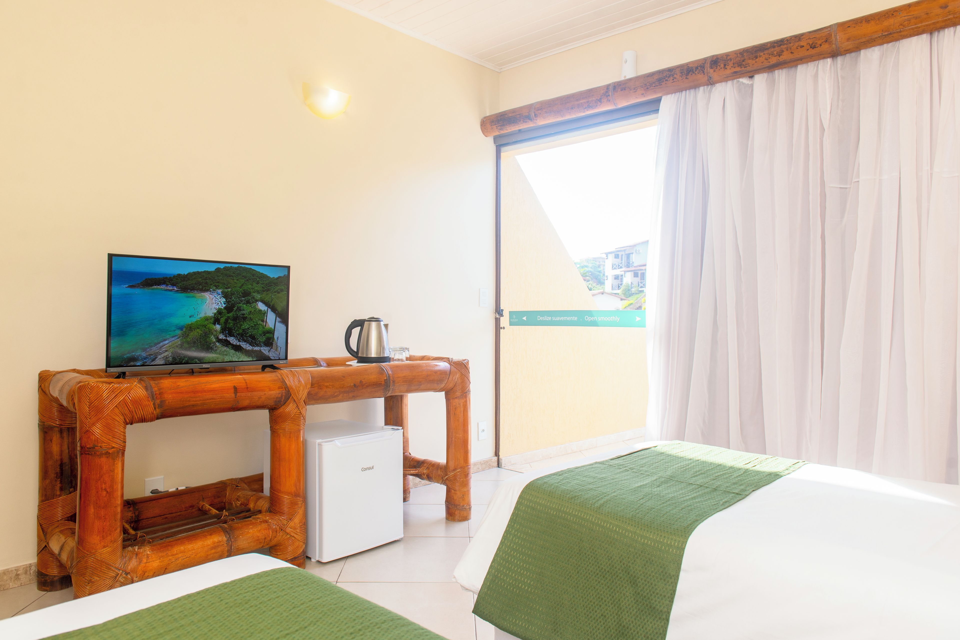 A hotel room with a bed, a small desk with a television, a coffee maker, and a view of a beach through the window.