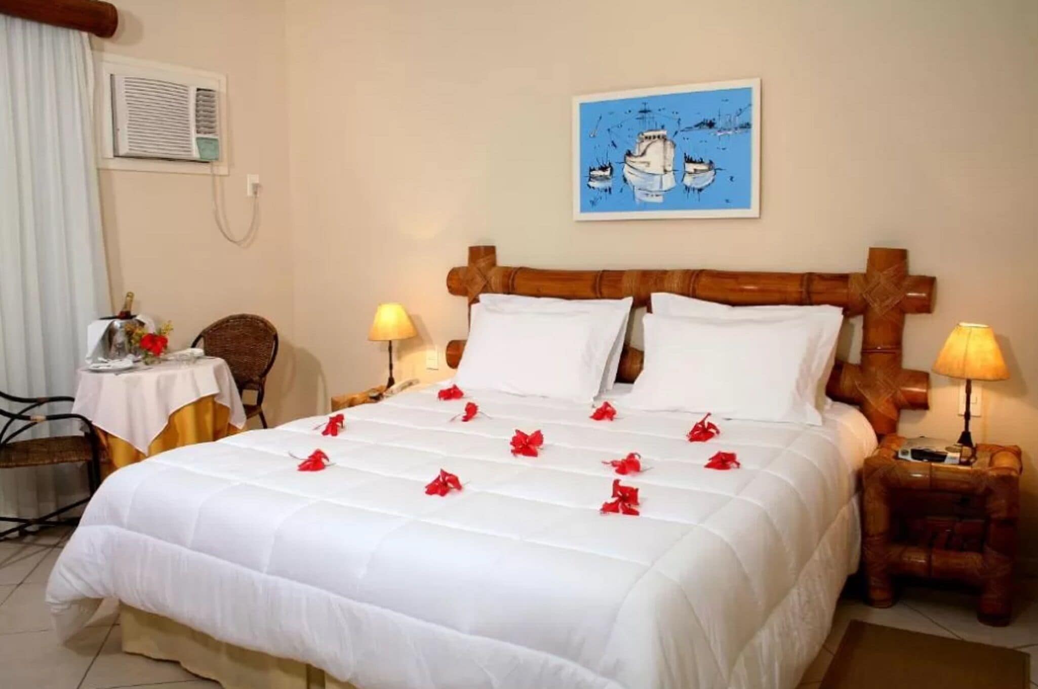 A neatly made bed with white bedding and red flowers, a wooden headboard, a small table with a lamp, and a chair.