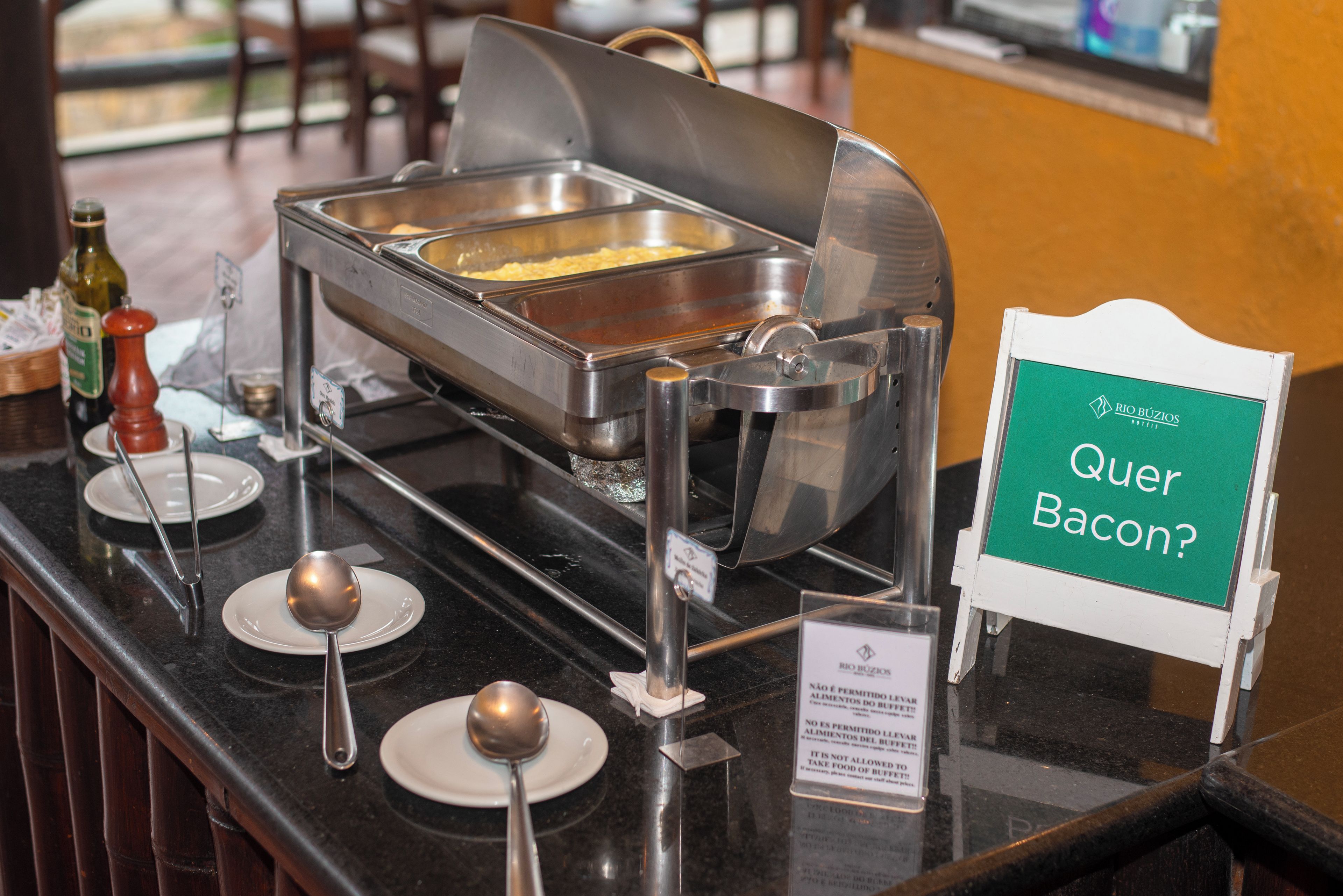 A buffet setup with a sign asking 'Quer Bacon?' indicating a bacon option.