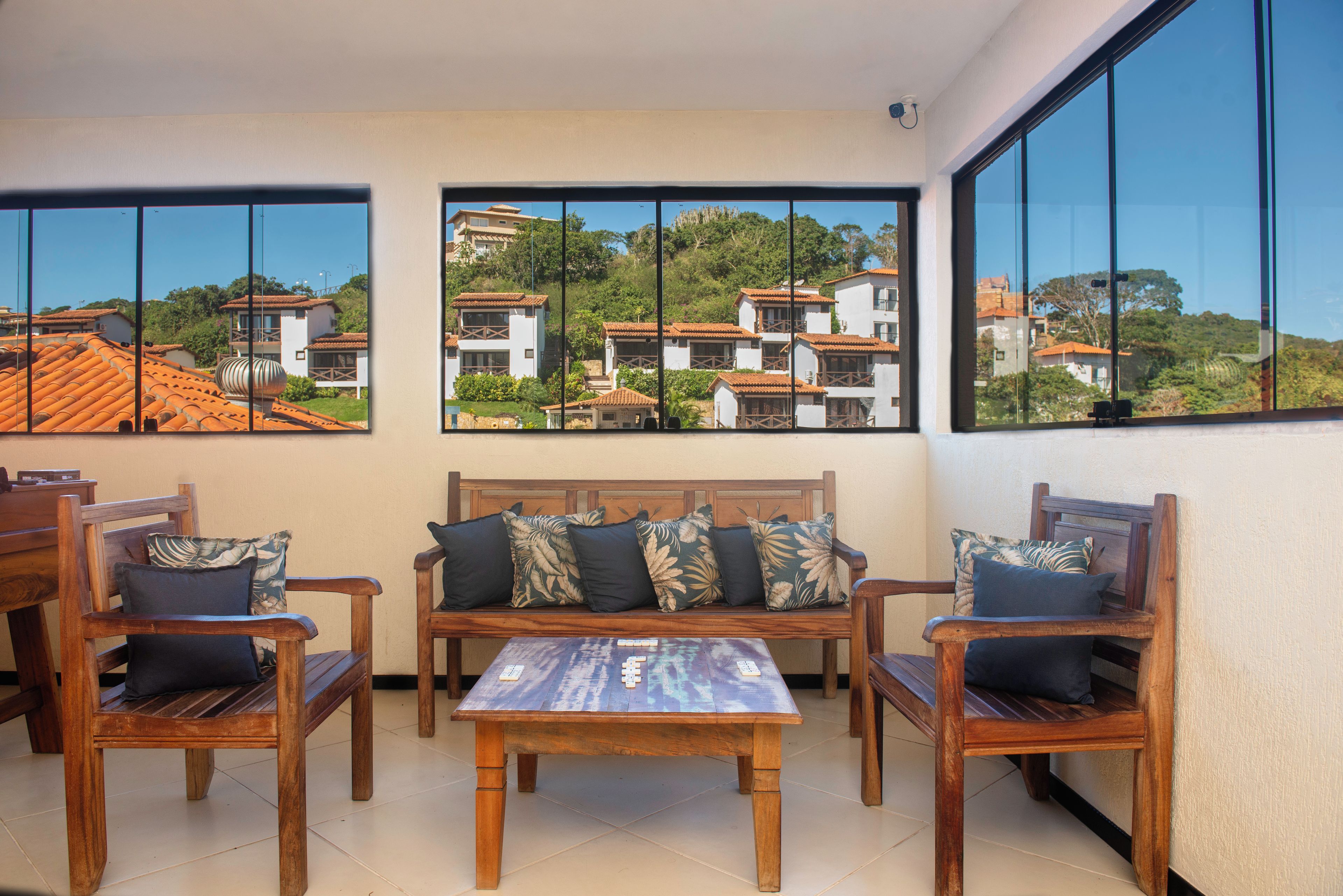 A sunlit room with a wooden sofa, two chairs, and a coffee table. Large windows offer views of buildings and greenery.