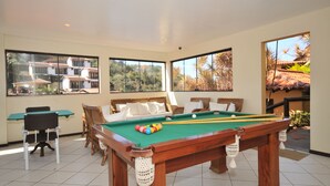 Una mesa de billar en una habitación con ventanas amplias, muebles de mimbre y vistas a un edificio y árboles.