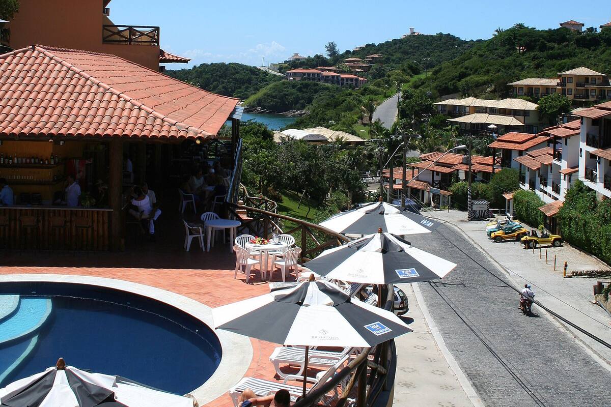 Un área junto a la piscina con sillones de descanso y sombrillas, un restaurante con terraza y vistas a una colina con edificios.