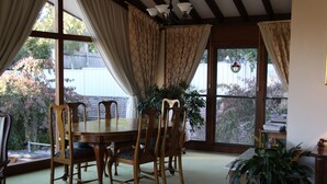 A dining area with a wooden table and chairs, large windows with curtains, and a potted plant. - Bathurst Heights B&B (Bathurst)