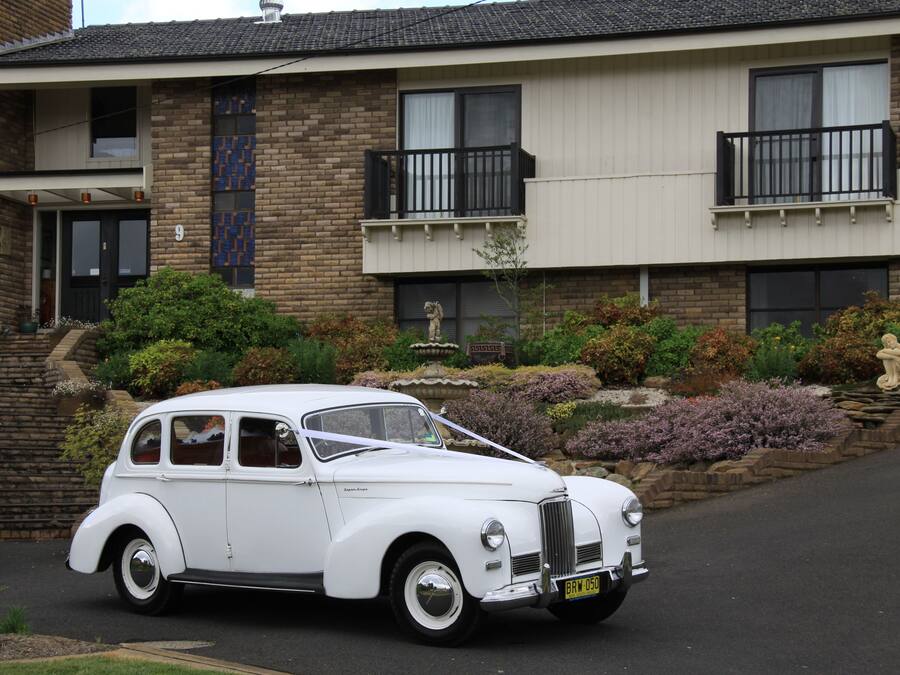 A white vintage car parked in front of a two-story house with a brick facade, a balcony, and a well-maintained garden with various plants and a statue.