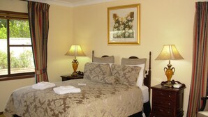 A neatly made bed with a floral bedspread, two bedside lamps, a framed floral artwork on the wall, and a window with curtains. - Bathurst Heights B&B (Bathurst)