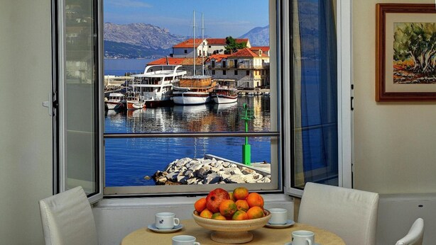 Una mesa de comedor con un cuenco de fruta y tazas, una ventana con vistas a barcos y edificios junto al agua.