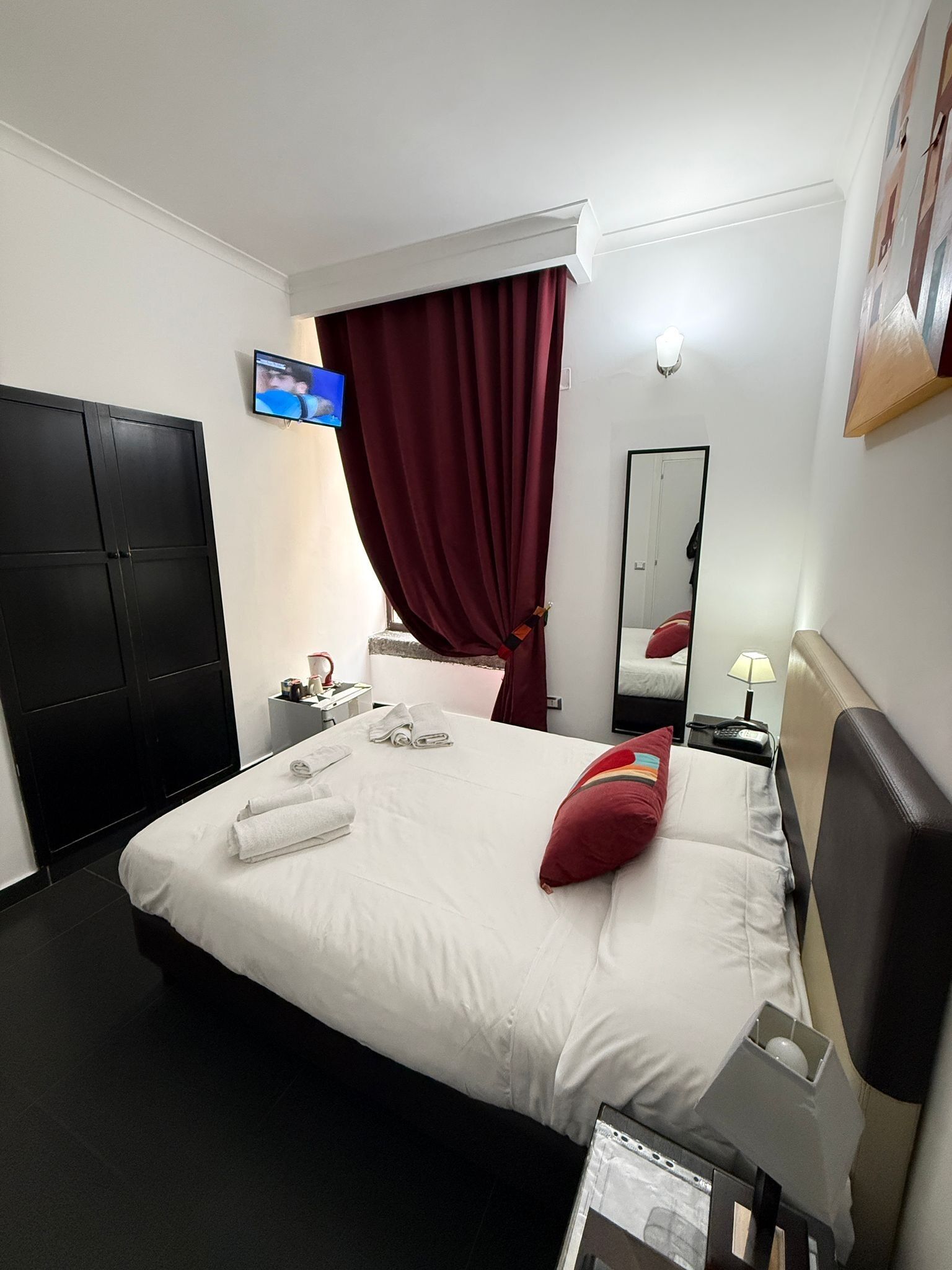 Quarto de hotel com cama, televisão, escrivaninha, cadeira, espelho e guarda-roupa.