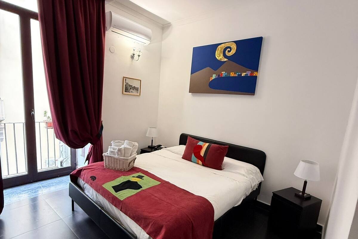 A modern hotel room with a large bed, a balcony with red curtains, a cityscape painting, and a small table with a lamp.