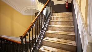 Un escalier en marbre, doté d’une rampe en bois et de balustres noires, mène à un palier où se trouvent un extincteur et un tableau accroché au mur.