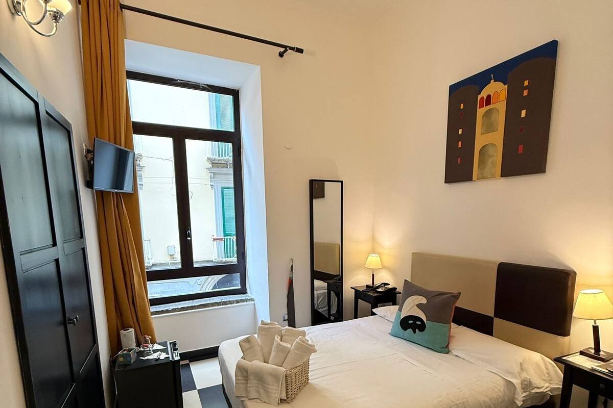 A hotel room with a checkered floor, a bed with white linens, a window with curtains, a flat-screen TV, a bedside lamp, and a painting on the wall.