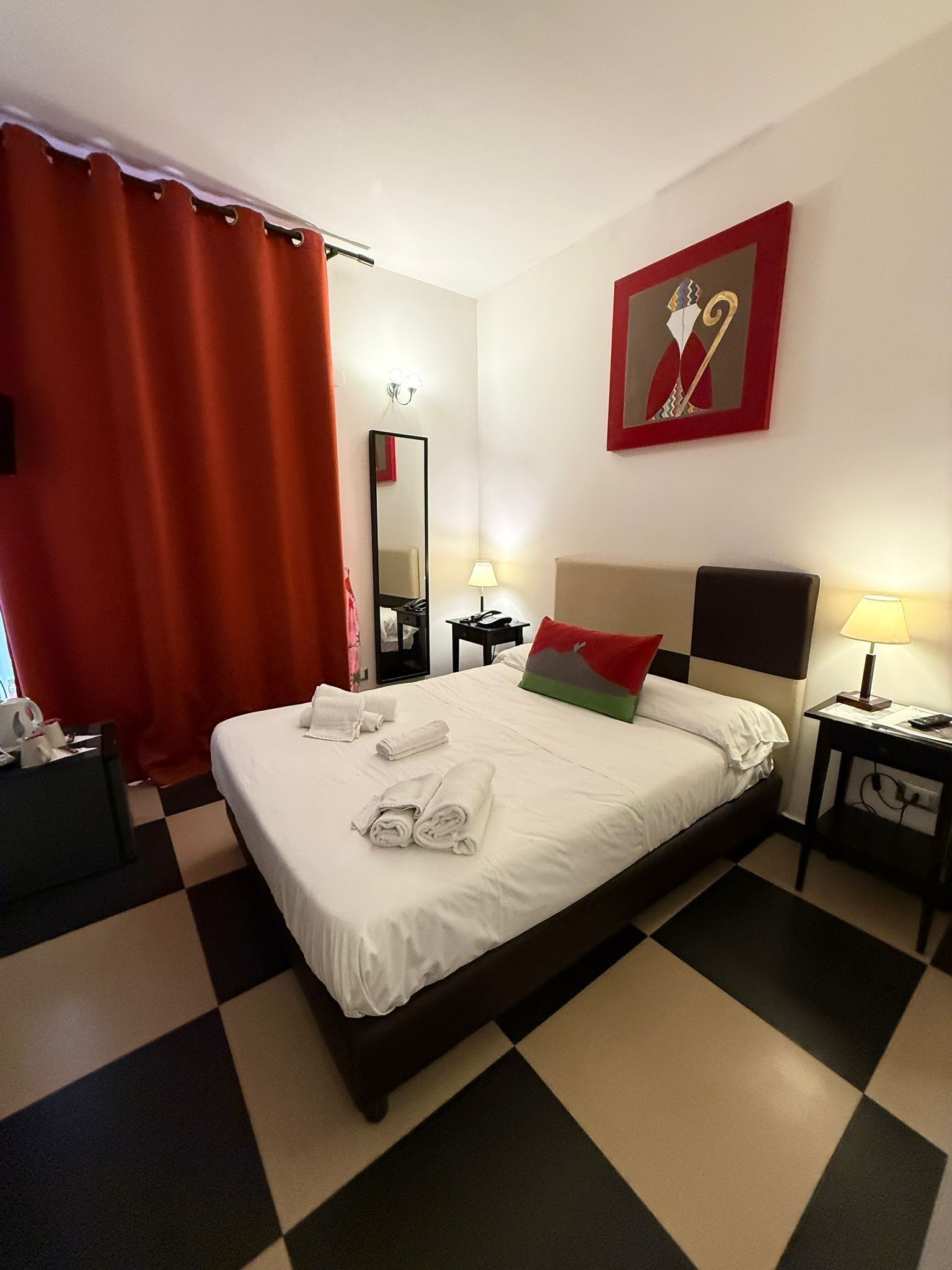 A hotel room with a checkered floor, a bed with white linens, a red curtain, a mirror, two bedside tables with lamps, and a painting on the wall.