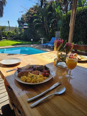 A breakfast table with a plate of food, a glass of orange juice, and a view of a swimming pool and garden. - 113 On Robberg (Plettenberg Bay)