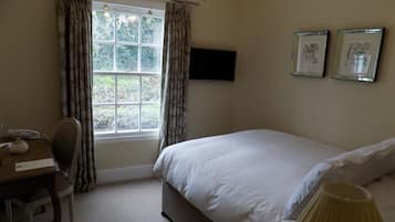 Un dormitorio con cama, escritorio, silla, ventana con cortinas, televisor de pantalla plana y dos cuadros en la pared.