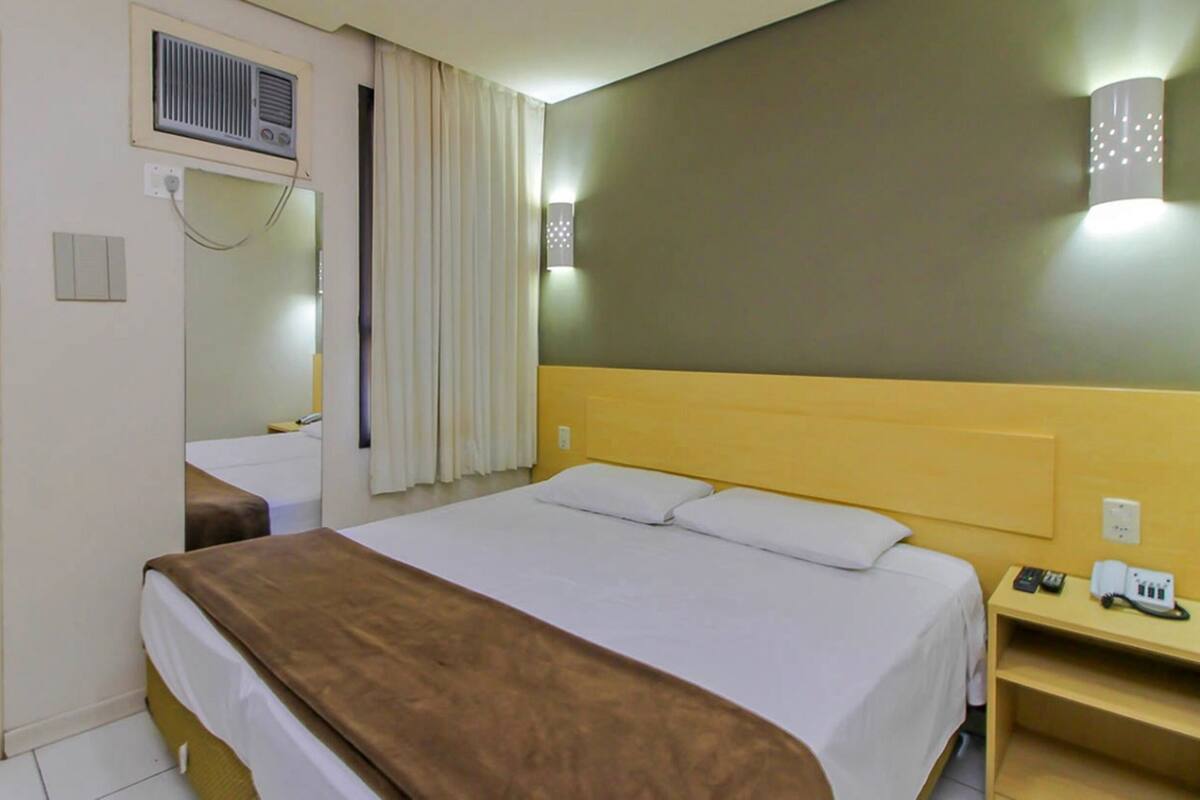 A hotel room with a bed, bedside table, lamp, and a wall-mounted air conditioning unit.