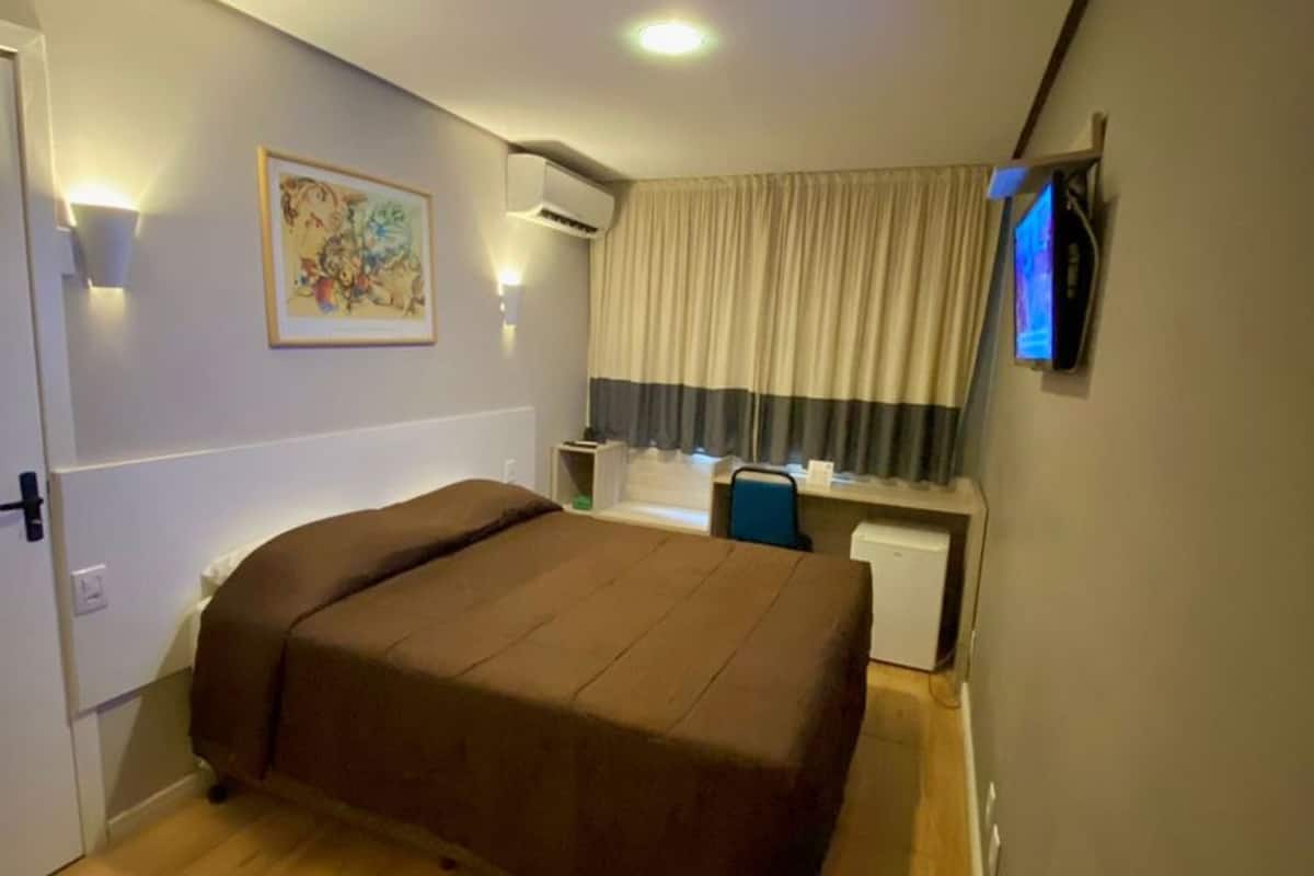 A bedroom with a bed, a desk, a chair, an air conditioning unit, and a wall-mounted TV.